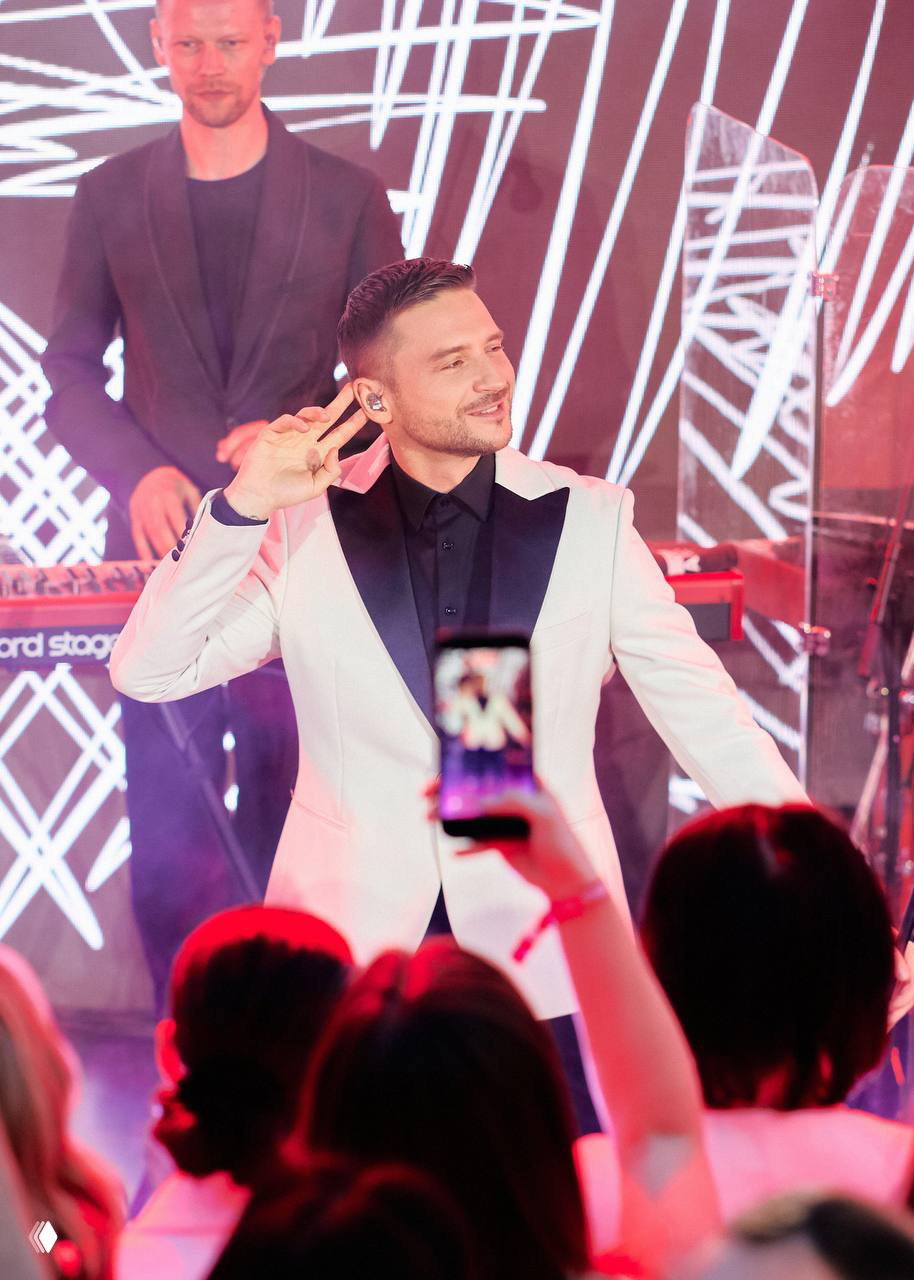 Сергей Лазарев на сцене Marie Claire Blogger Party 2026 во время выступления: певец в белом пиджаке энергично взаимодействует с публикой и камерой.