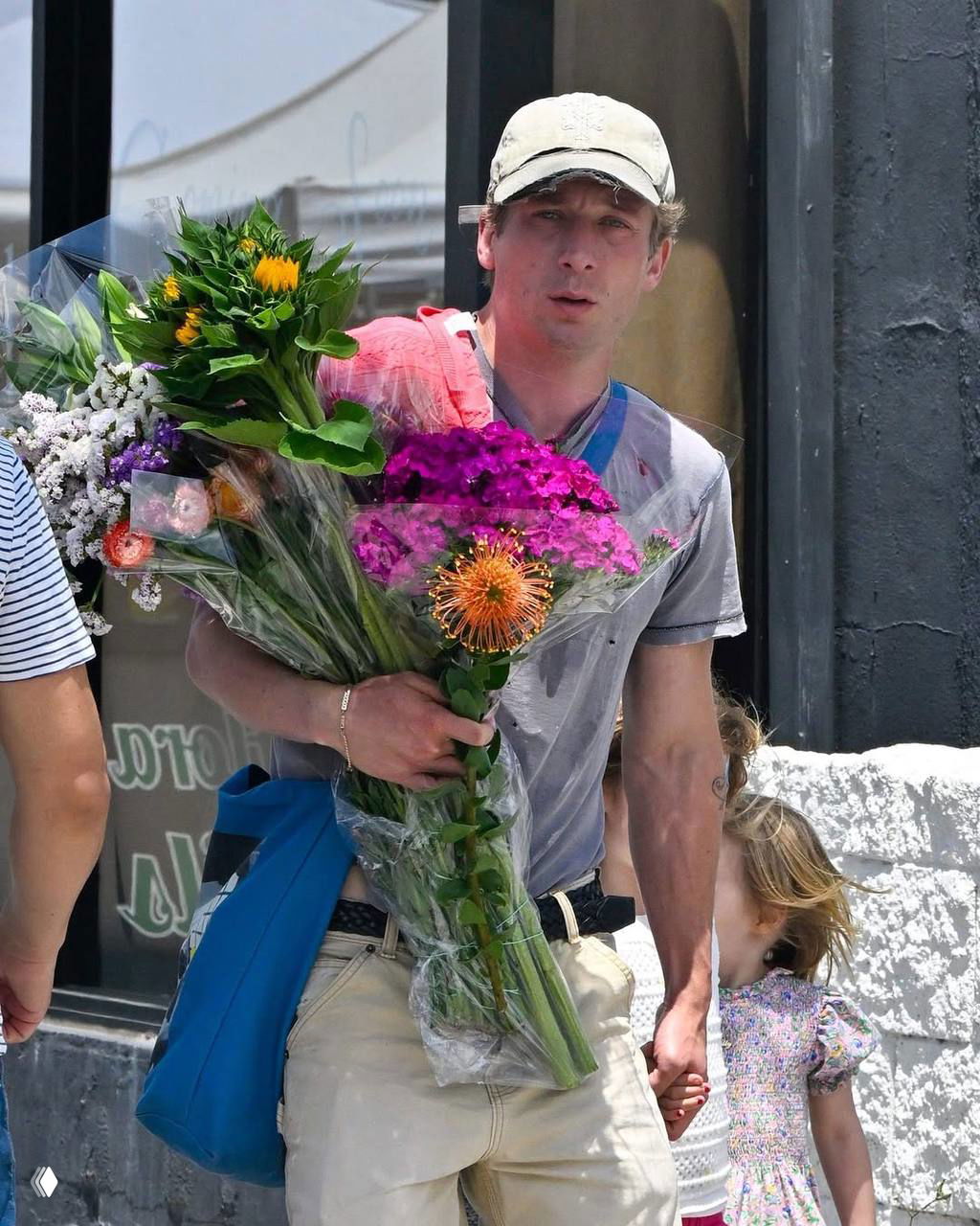 Мужчина несёт большой букет ярких цветов и держит за руку маленького ребёнка; кадр семейной прогулки по городу в тёплый день, естественный свет.