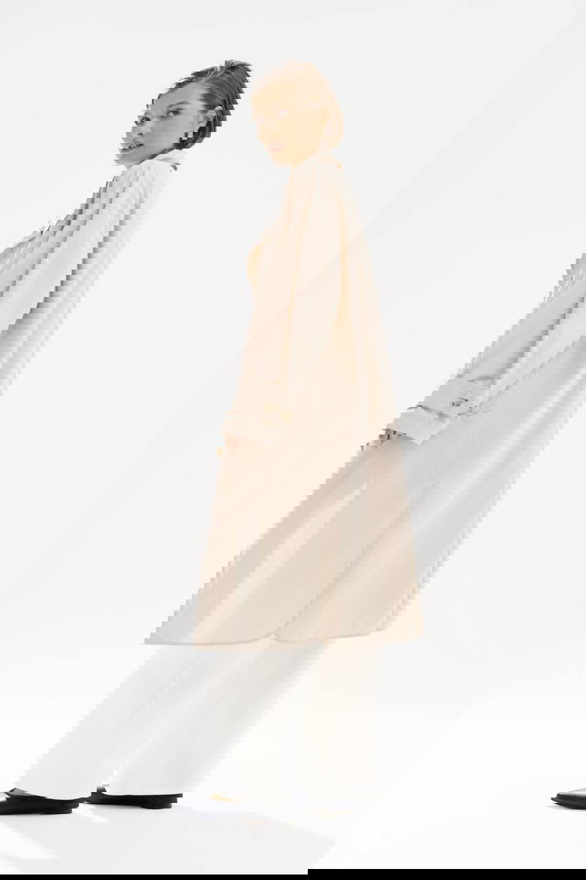 Фотография коллекции Nelva: профиль модели в кремовом тренче и белых широких брюках на минималистичном фоне — элегантный городской лук.