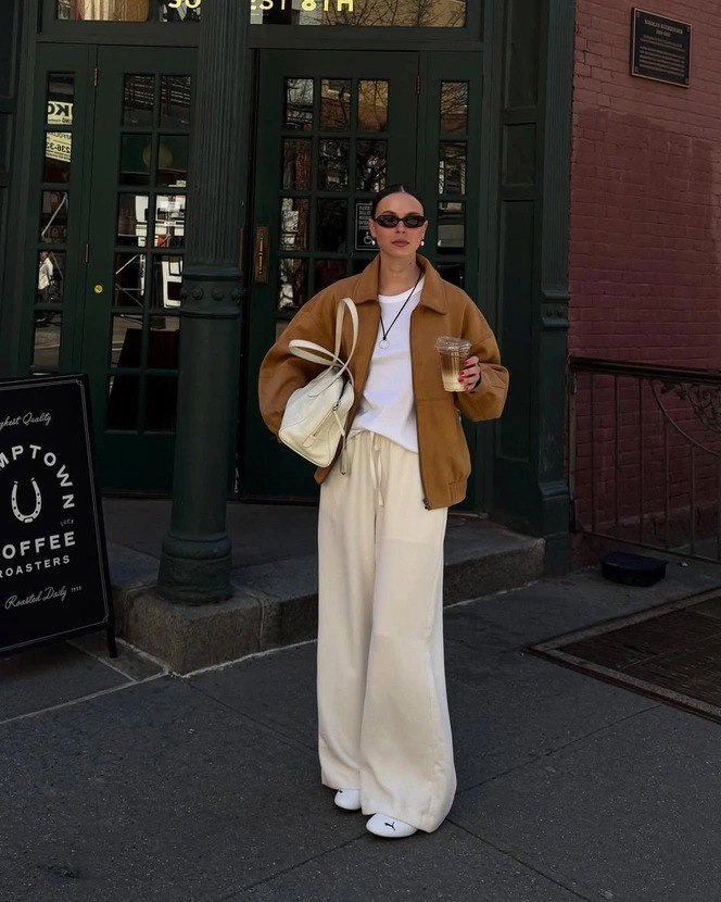 Уличный образ: широкие кремово‑белые брюки, коричневая куртка‑бомбер, белая сумка и кроссовки; девушка идёт по городскому тротуару с напитком в руках.