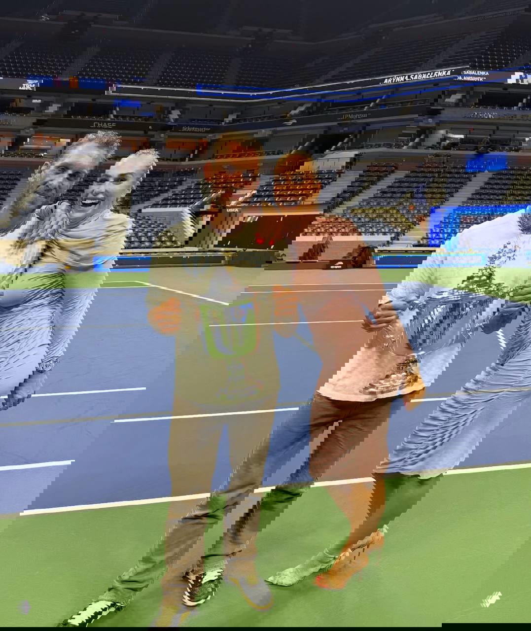 Георгиос Франгулис и Арина Соболенко стоят на корте с крупным трофеем, вокруг пустые трибуны и спортивная арена после церемонии.