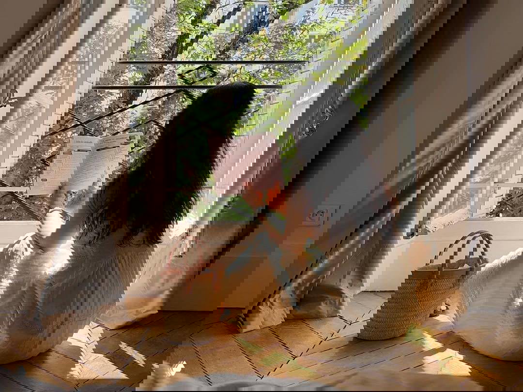 Женщина в светлом платье сидит у открытого окна и читает: деревянный паркет, сумка рядом, мягкий дневной свет и вид на зеленые ветви за окном.