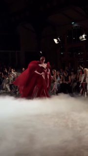 Два силуэта моделей в тёмных и красных нарядах Iris Van Herpen на подиуме; динамичный кадр шоу, дым и сильные сценические эффекты.