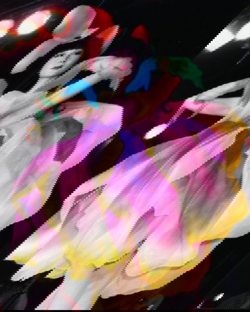 Модель в ярком эффектном платье‑цветке Mugler 1982 на тёмном фоне сцены: крупные лепестки с розово‑жёлто‑фиолетовым градиентом.