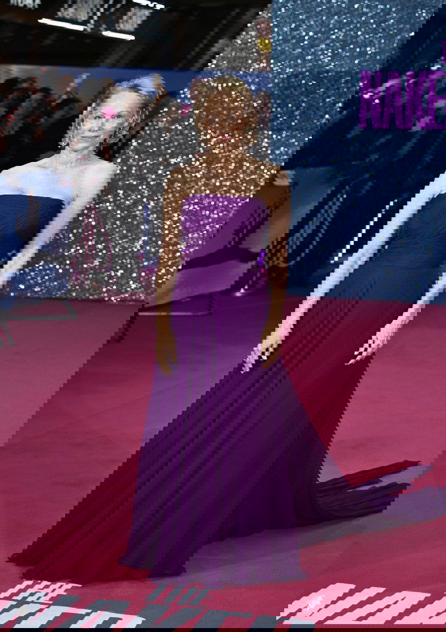 Элегантная женщина в сдержанном фиолетовом платье в полный рост на красной дорожке премьеры, спокойный силуэт и длинная юбка на фоне фотоколла.