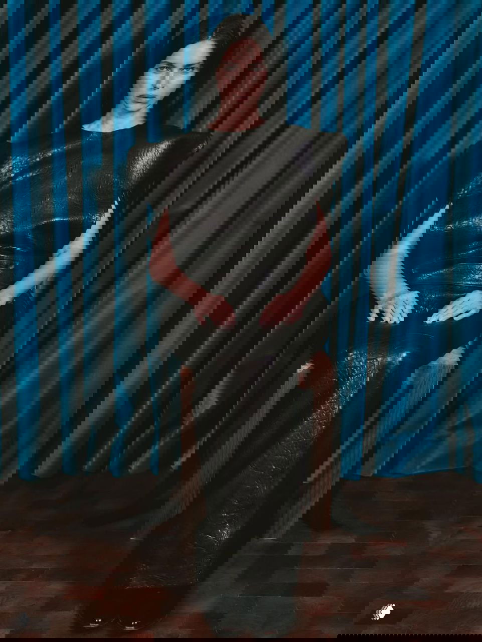 Модель сидит в структурированном чёрном кожаном платье на стуле на фоне плотной синей бархатной шторы, холодный и строгий взгляд в камеру.