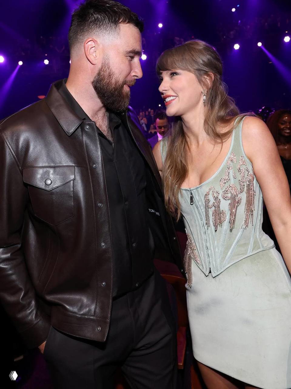 Трэвис Келси и Тейлор Свифт общаются в зале церемонии, певица улыбается в тусклом фиолетовом свете сцены, пара в вечерних нарядах.