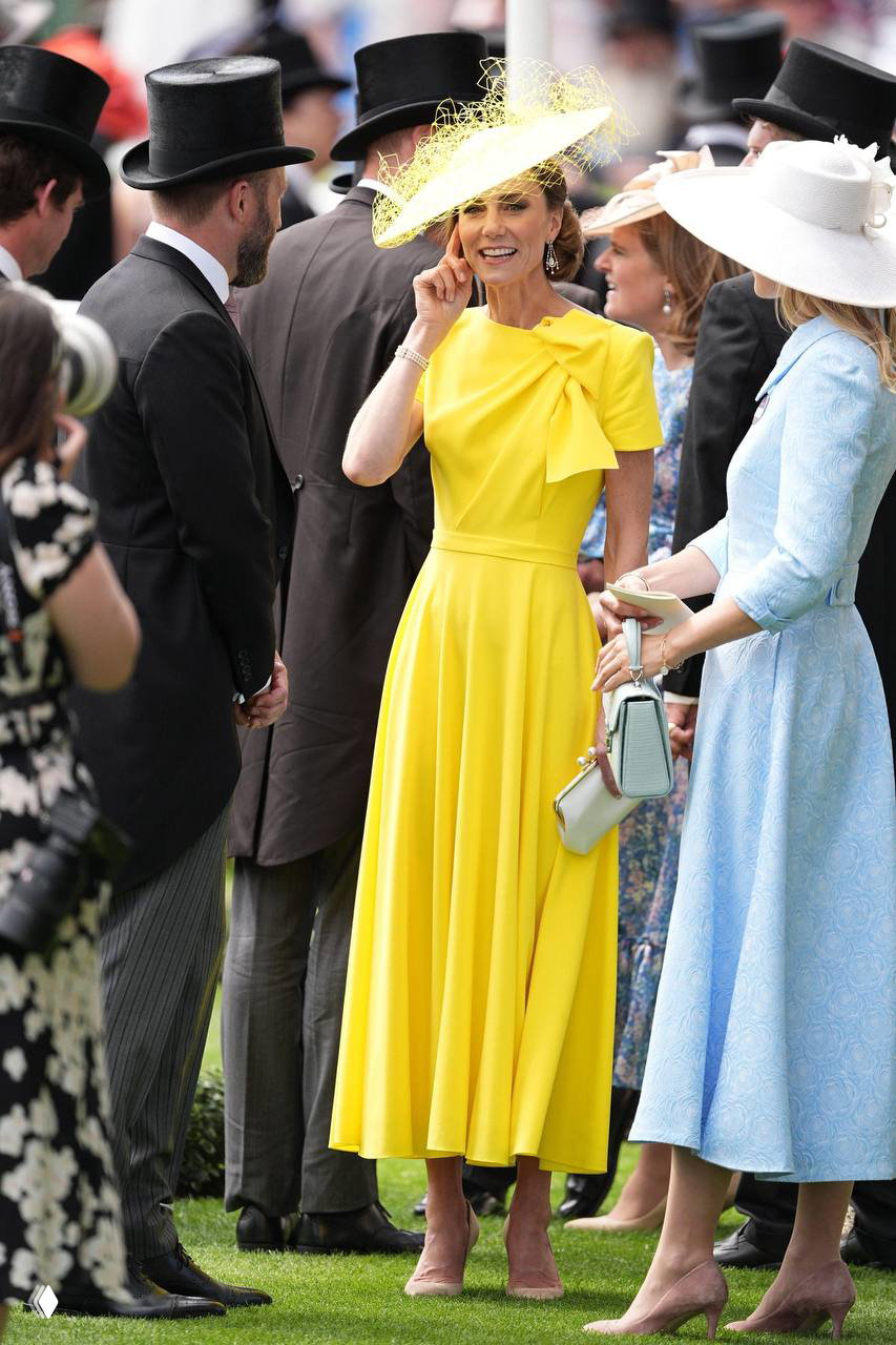 Женщина в жёлтом миди‑платье и шляпе стоит на лужайке Royal Ascot среди гостей в цилиндрах; видна сумочка, туфли и окружение праздничного мероприятия
