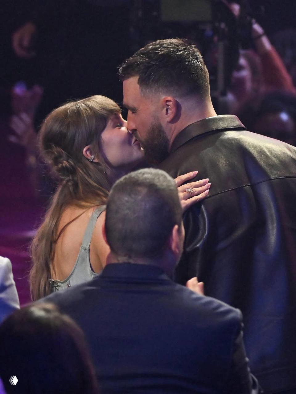 Тейлор Свифт целует Трэвиса Келси в зале награждения; видны спины сидящих людей и фрагменты сцены, intimate-кадр в полутёмном помещении.