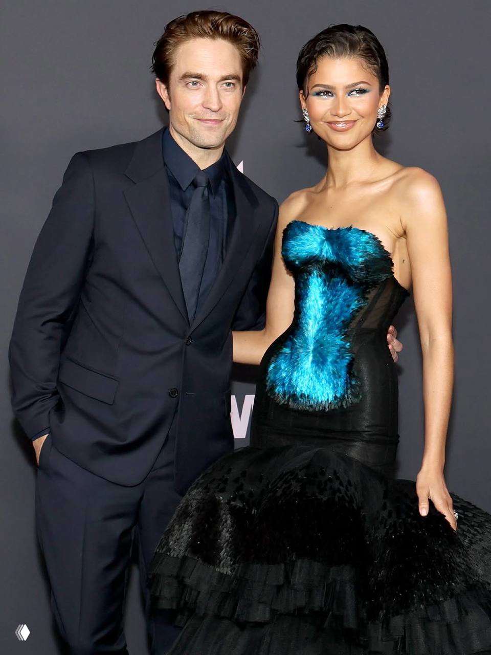 Пара на красной дорожке: женщина в платье с перьями и мужчина в классическом тёмном костюме позируют для фотографов.