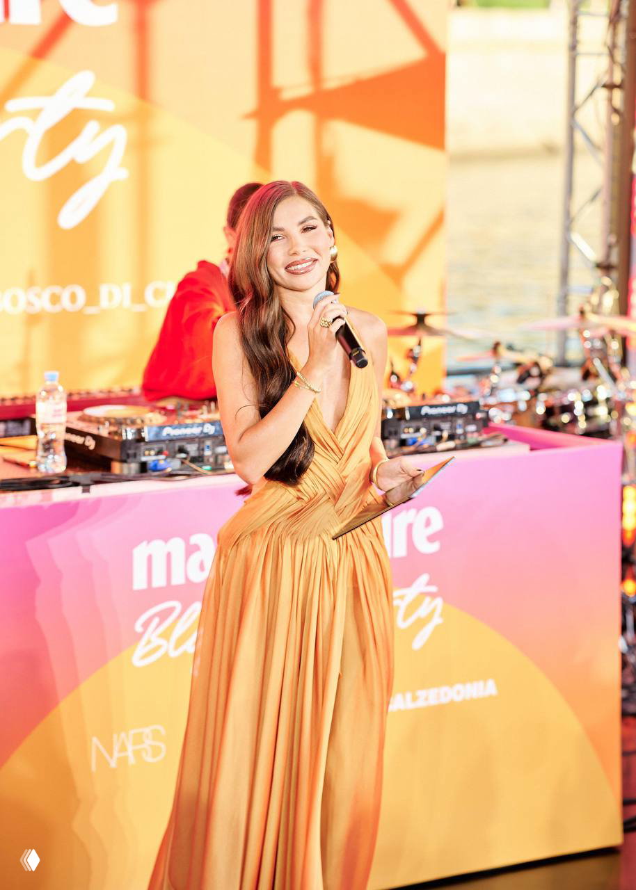 Ведущая мероприятия в золотом платье на сцене Marie Claire Blogger Party 2026: микрофон, диджейский пульт и тёплая сценическая подсветка.