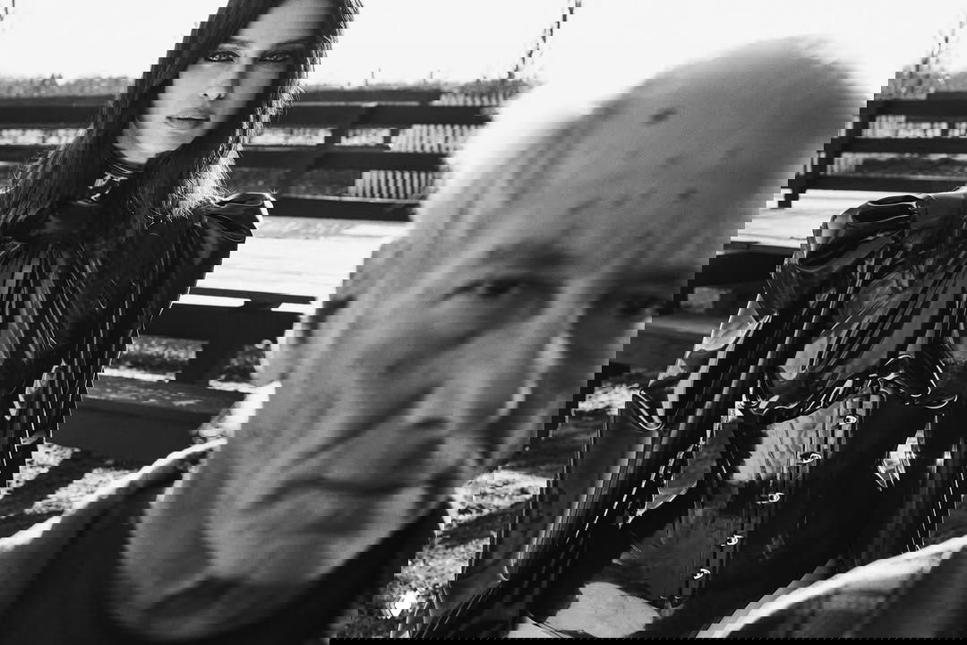 Чёрно-белый портрет: модель в глянцевом чёрном плаще и корсете на открытом воздухе, на переднем плане размыто лицом пожилого мужчины, режиссёрский приём кадра.