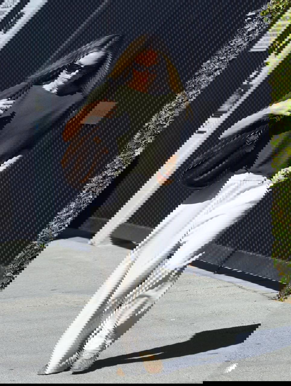 Днёвная уличная фотография: женщина в свободной оливковой футболке и белых широких брюках идёт по тротуару с большой сумкой, городской фон.