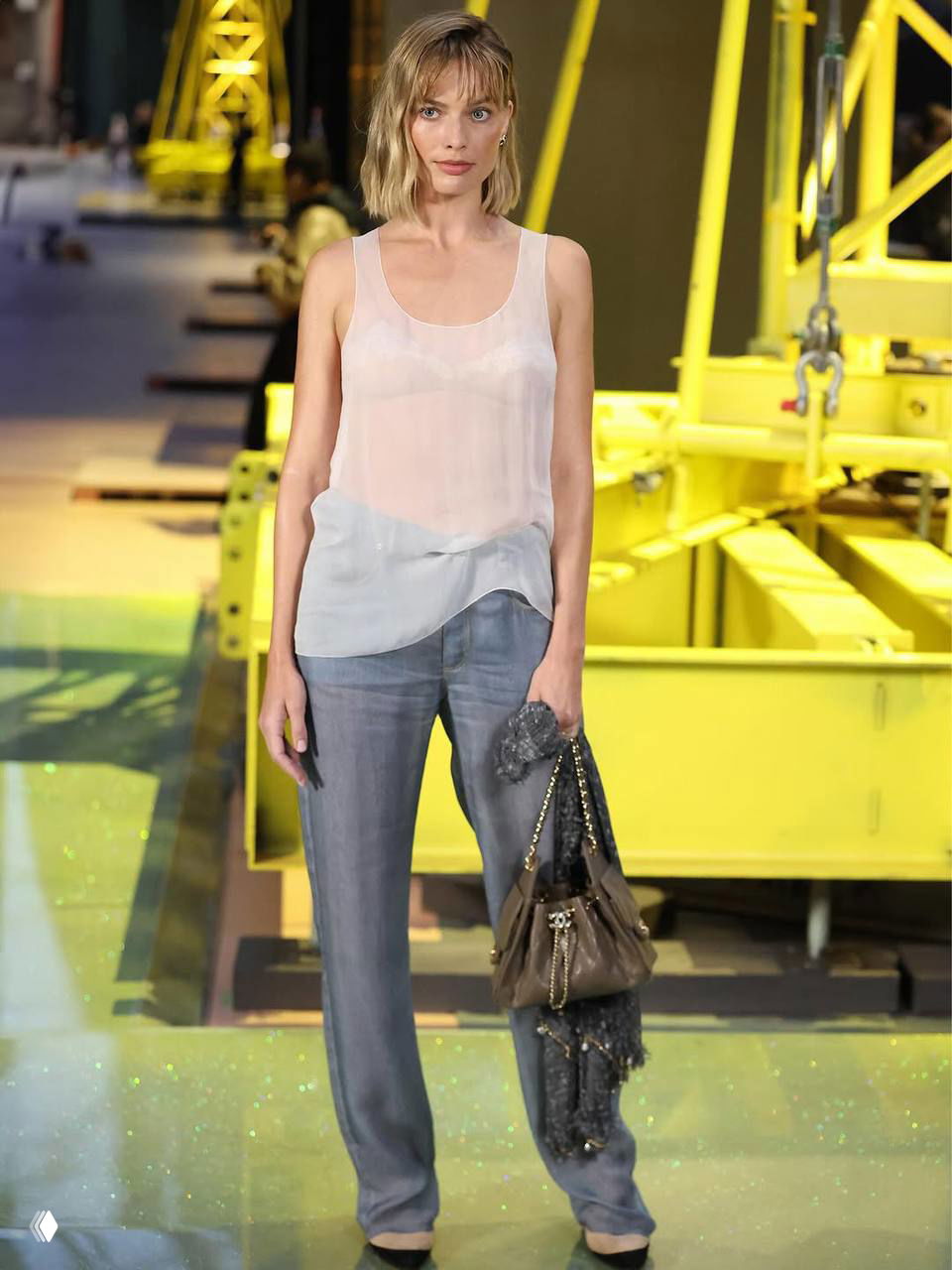 Марго Робби на показе Chanel в Париже — белая полупрозрачная майка, серые брюки и сумка на подиуме с желтыми декорациями.