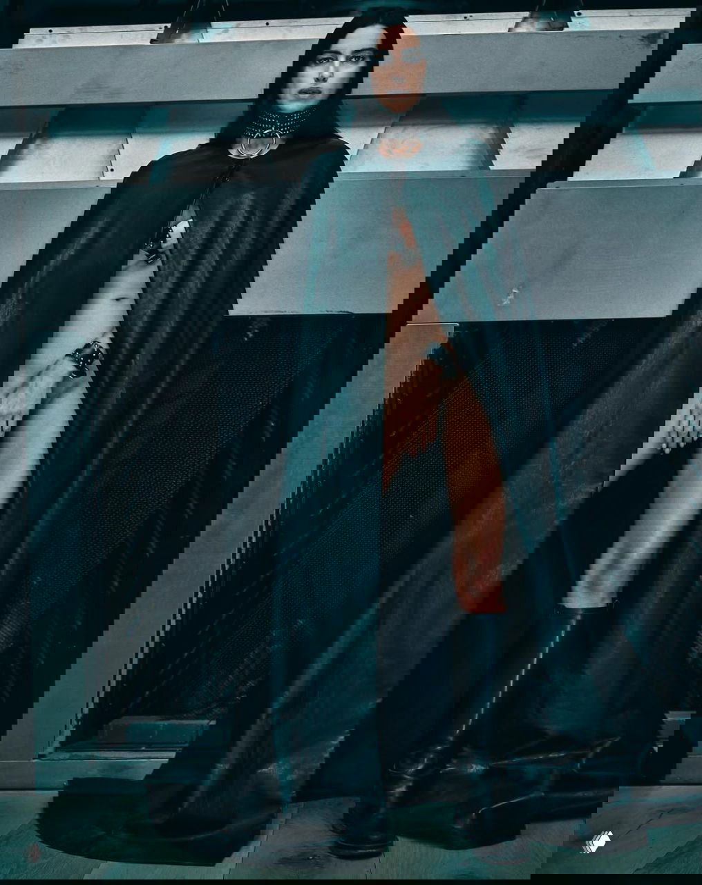 Модель в длинном чёрном плаще с открытым разрезом и массивным чокером, индустриальный интерьер и сильный светотеневой контраст в кадре.