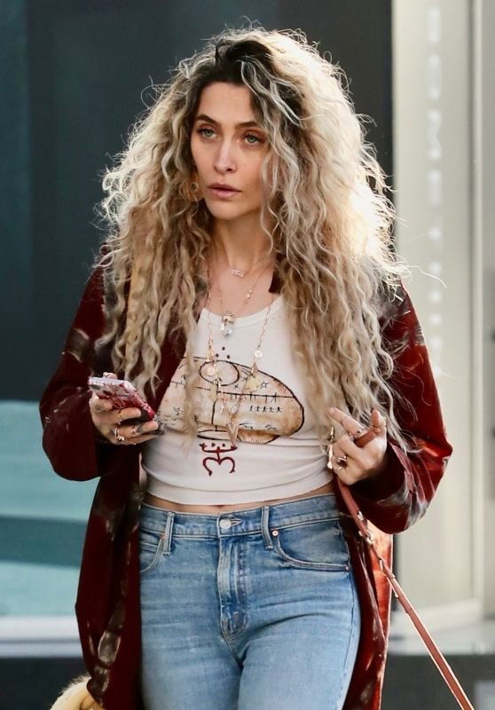 Крупный план: женщина с длинными вьющимися волосами в повседневном образе — джинсы и бордовая куртка на фоне городской прогулки, естественное освещение.