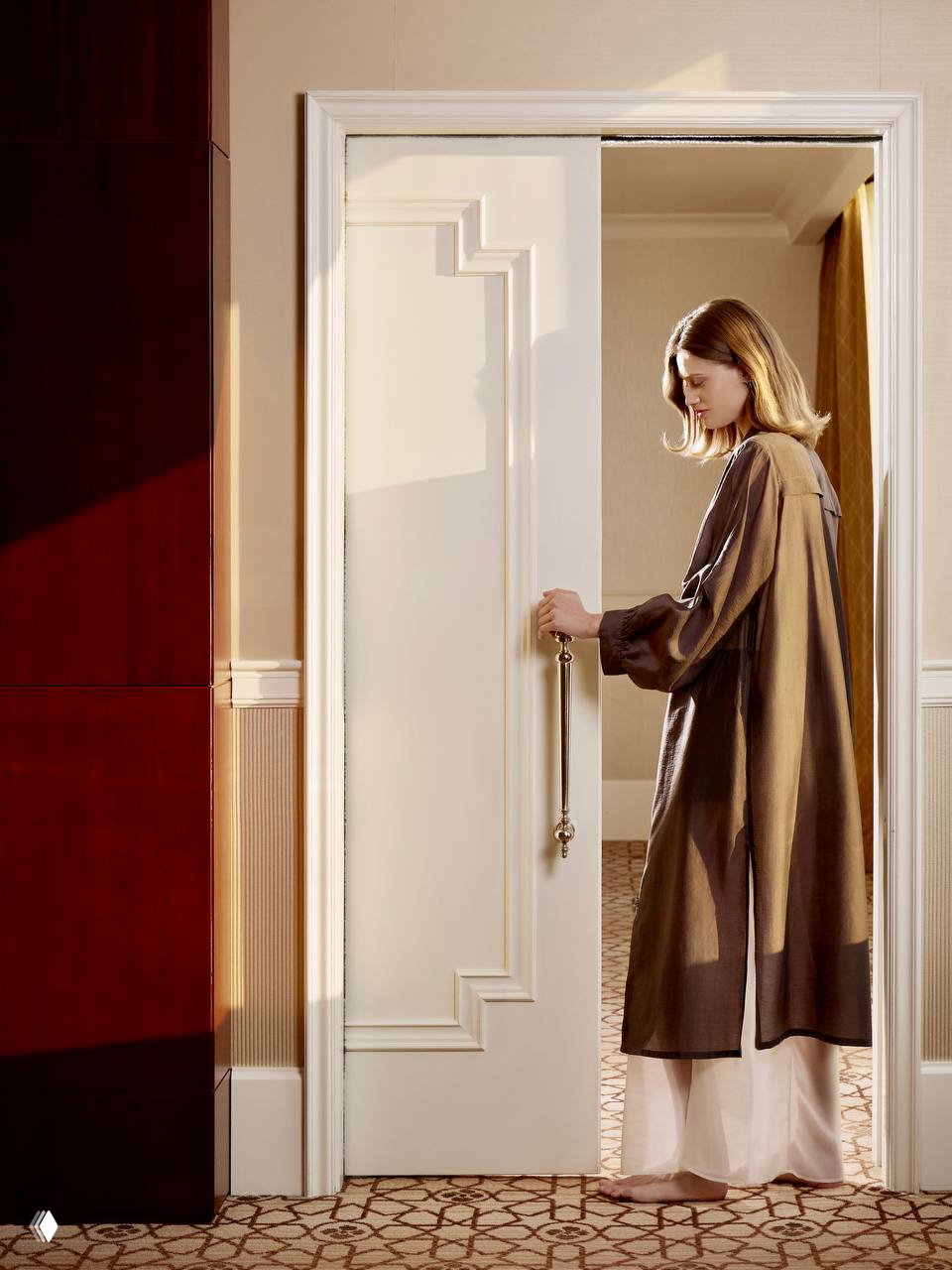 Модель в длинном полупрозрачном халате стоит у классической двери гостиничного номера, мягкий свет и внимание к силуэту и архитектуре интерьера.
