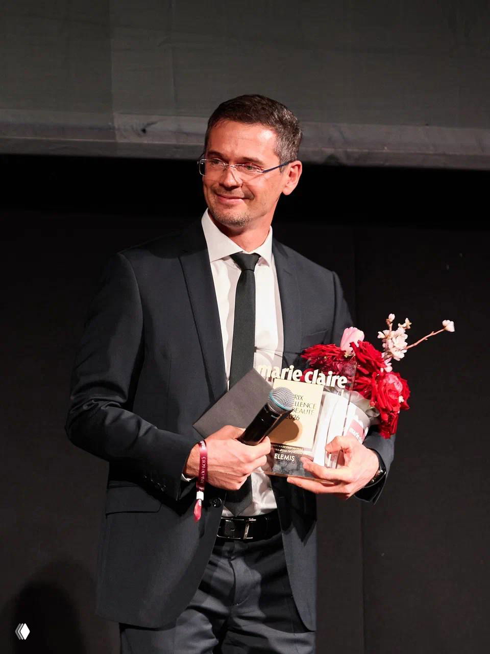 Мужчина в костюме держит награду Marie Claire Prix d'Excellence de la Beauté 2026 и букет цветов, сценический кадр с микрофоном и тёмным фоном.