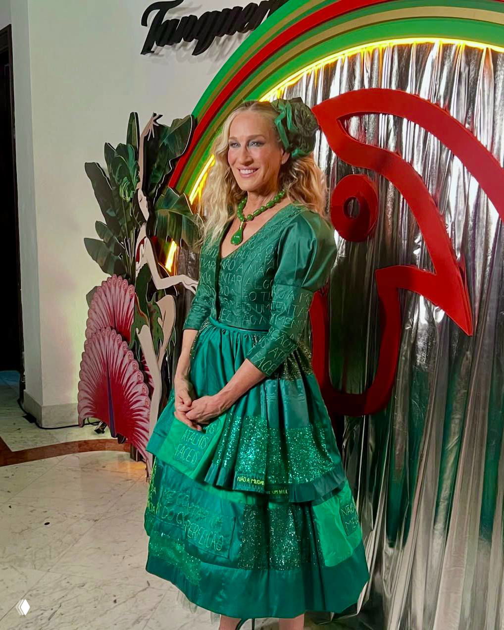 Сара Джессика Паркер в полный рост у инсталляции Baile da Arara; изумрудное платье Изабелы Капето и аккуратная причёска с декоративным цветком.