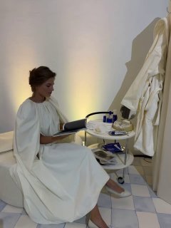 Женщина в элегантном белом платье сидит рядом со столиком с косметикой и аксессуарами; кадр бекстейджа и подготовки образов для показа YUDASHKIN.