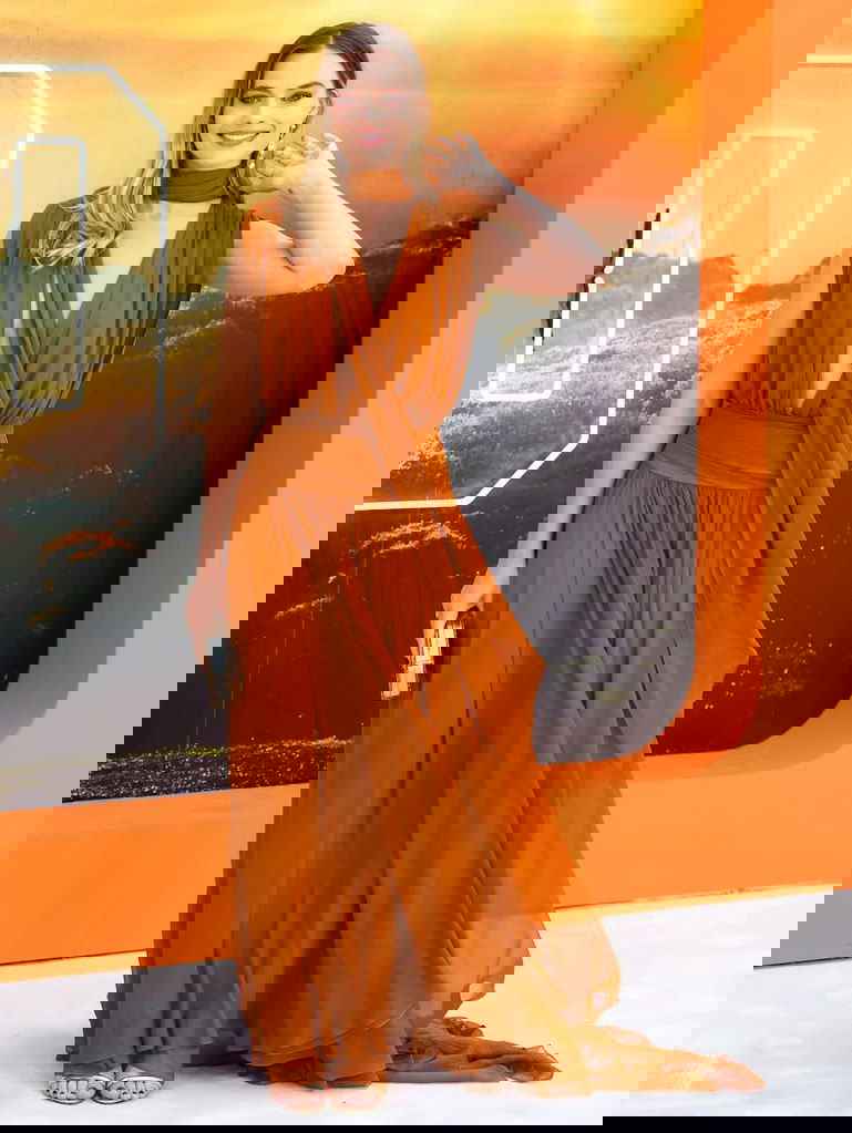 Женщина в струящемся оранжевом платье с глубоким декольте и поясом улыбается на фоне оранжевой декорации, в руках клатч