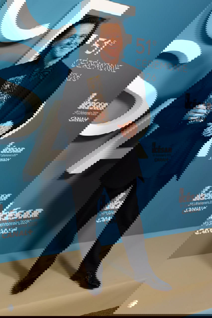 Джим Керри позирует с наградой у фотозоны 51-й церемонии «Сезар», полный рост в чёрном костюме на светлом фоне.