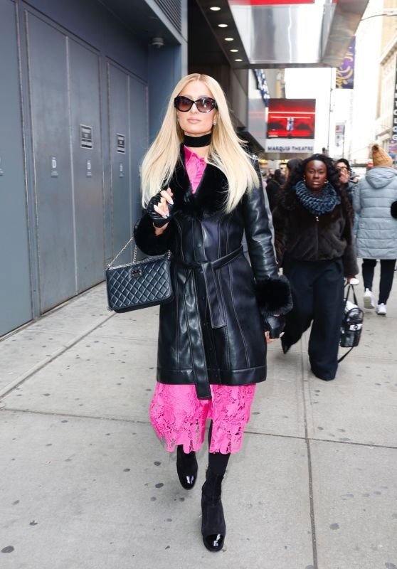 Женщина в длинном чёрном кожаном пальто и розовом платье идёт по тротуару в городской зимней обстановке, на заднем плане прохожие и вывески.