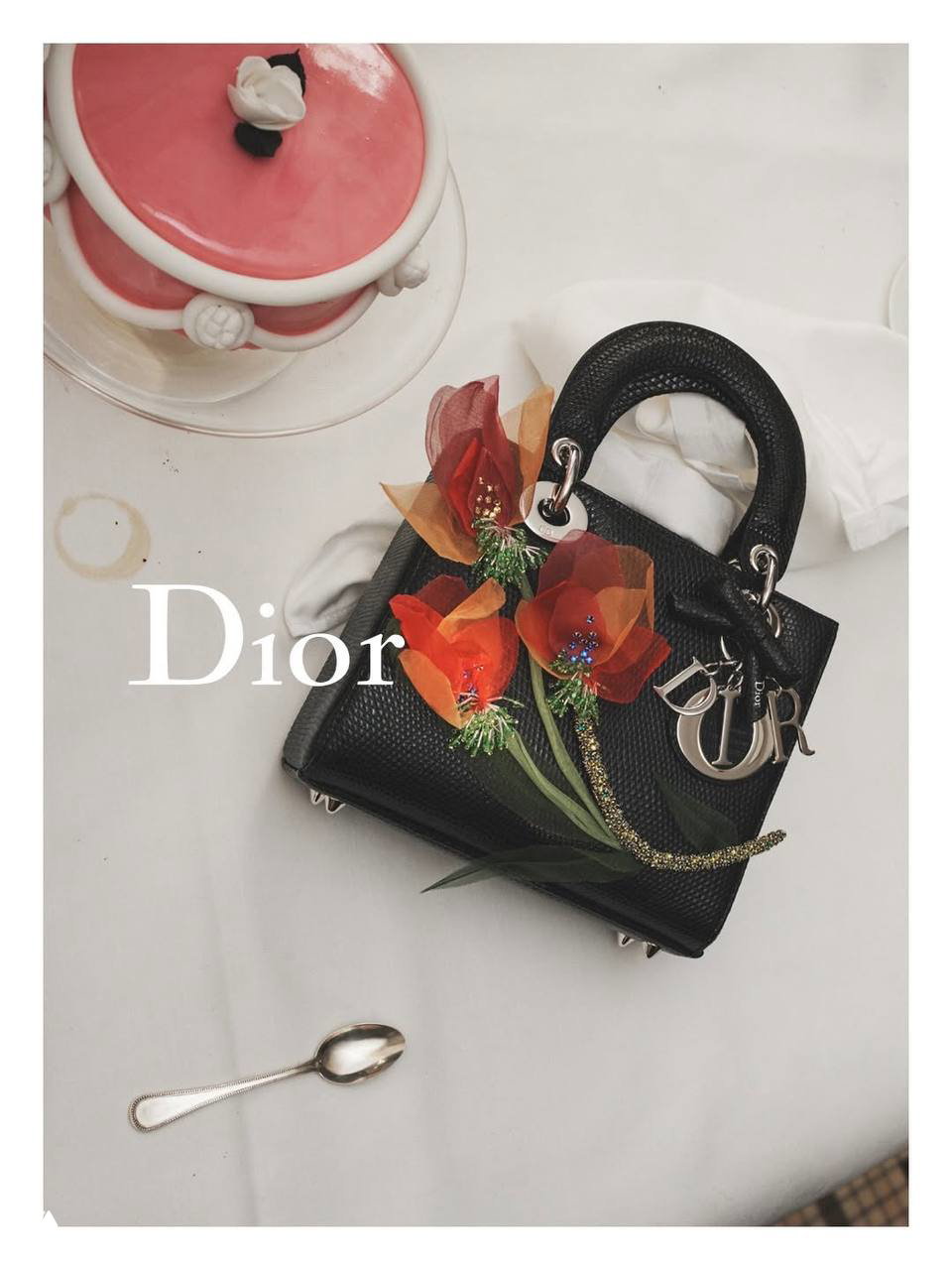 Миниатюрная сумка Dior на белой скатерти с цветочными декорациями и ложкой — рекламный натюрморт из кампании Café Society, фирменный логотип на кожаном аксессуаре хорошо различим.