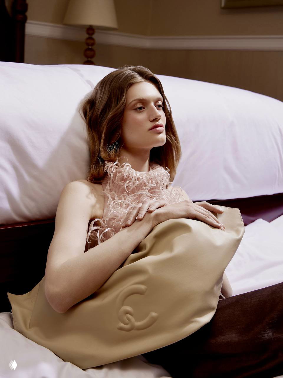 Крупный портрет модели на кровати в романтическом образе с текстурной блузой и большой сумкой, мягкое освещение подчёркивает детали съёмки.