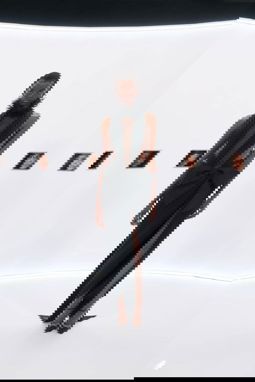 Стройная модель в чёрном платье с глубоким разрезом и бантом на талии на фоне минималистичной белой сцены с маленькими портретами.