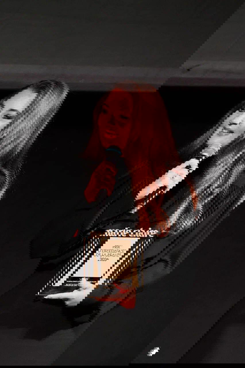 Женщина на сцене держит приз Marie Claire Prix d'Excellence de la Beauté 2026 и микрофон, портретный кадр с тёплым румяным светом и улыбкой.