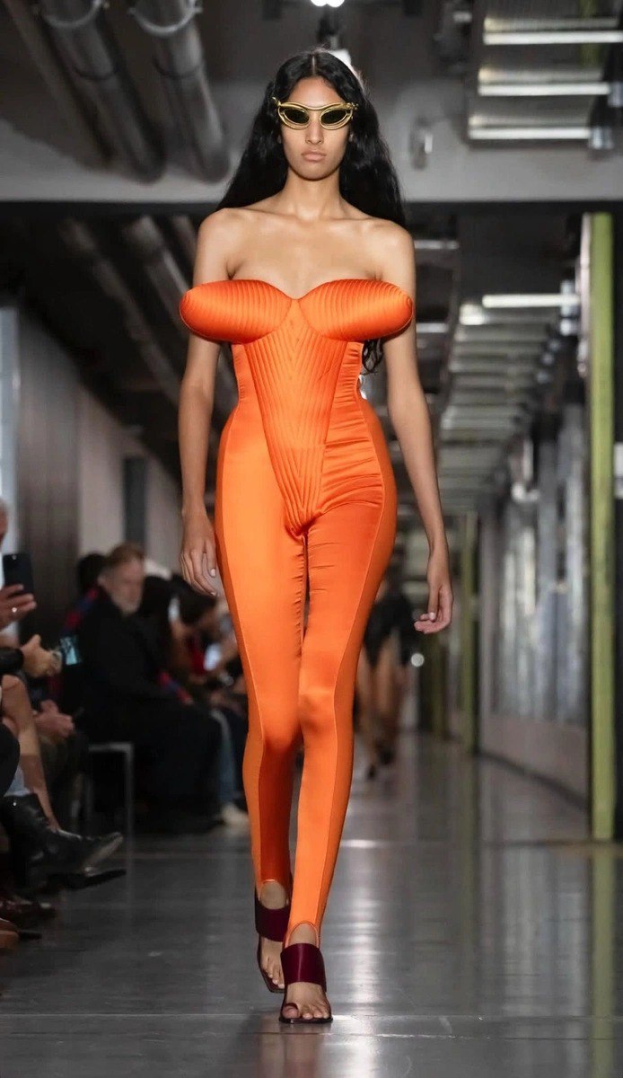 Модель в ярком оранжевом облегающем костюме с акцентом на бюсте — снимок с подиума показа коллекции Дюрана Лантинка для Jean Paul Gaultier.
