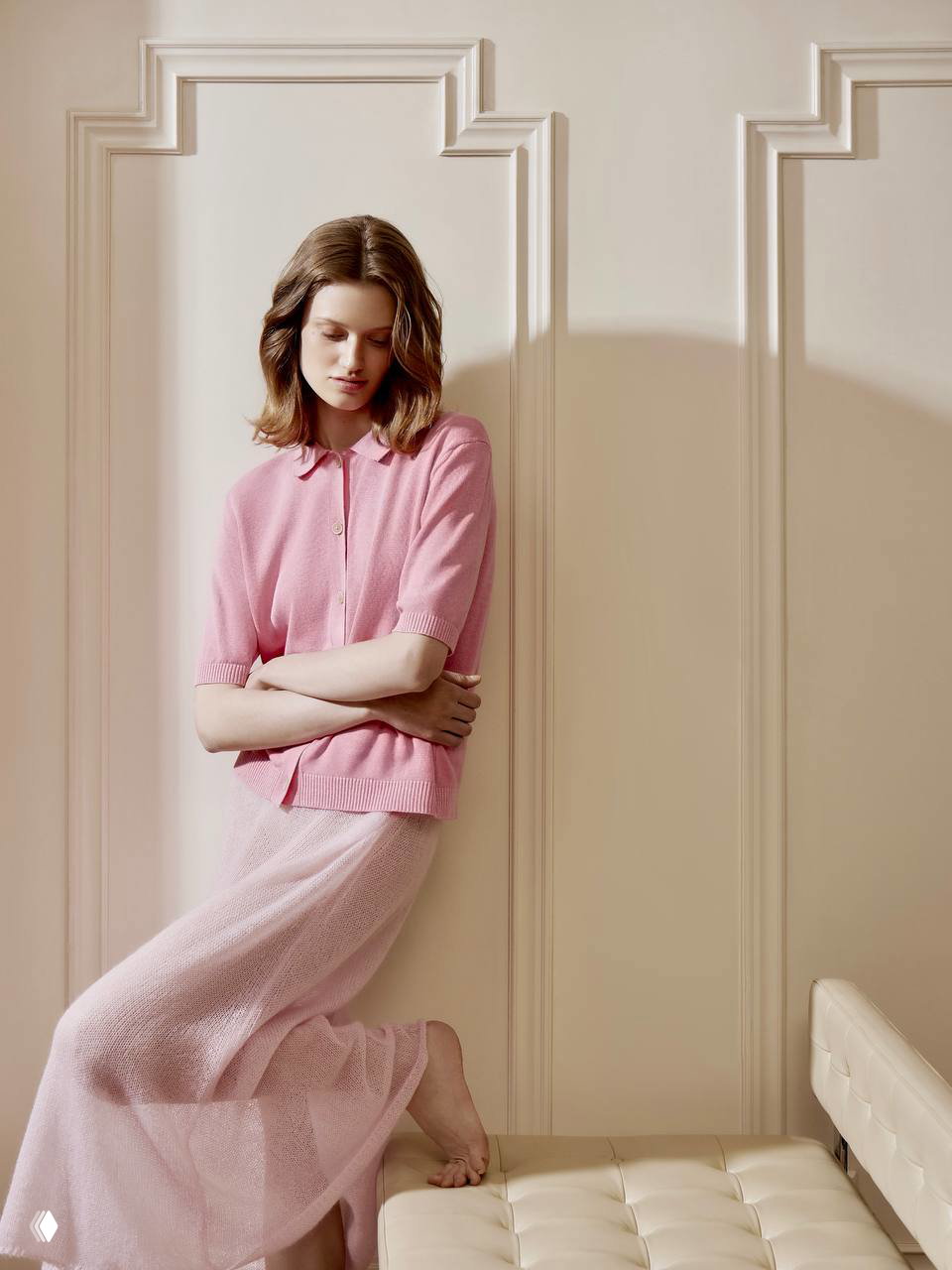 Модель в пастельно-розовом кардигане и лёгкой юбке позирует у классической декоративной панели в гостиничном номере, образ спокойный и элегантный.