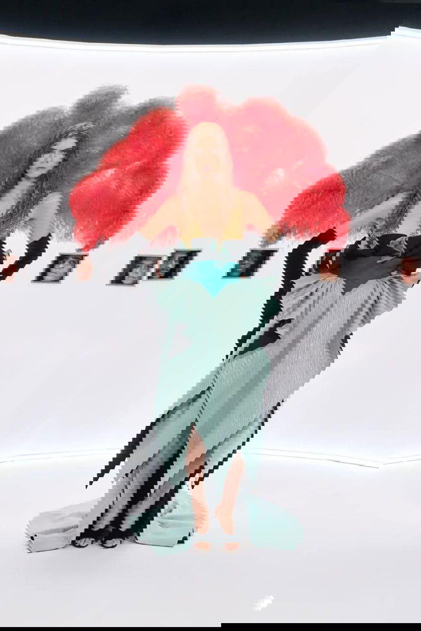 Шоу в сценографии: модель в экстравагантном костюме с большой красной перьевой деталью и многослойной юбкой, на белом круглом помосте.