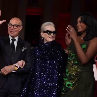 Кадр с тремя людьми на сцене премьеры: актёры аплодируют и улыбаются на фоне крупных красных декораций и вечерней подсветки.