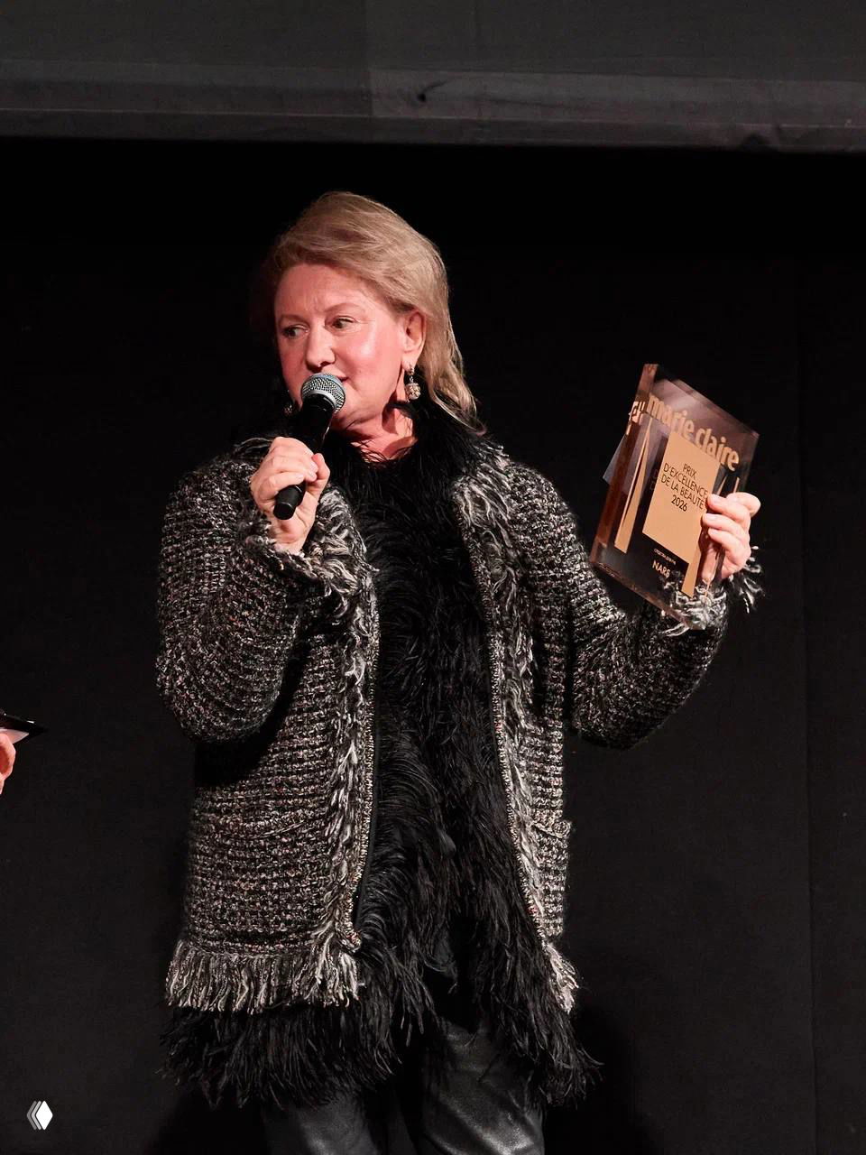 Пожилая женщина держит приз Marie Claire Prix d'Excellence de la Beauté 2026 и говорит в микрофон во время церемонии на тёмной сцене.