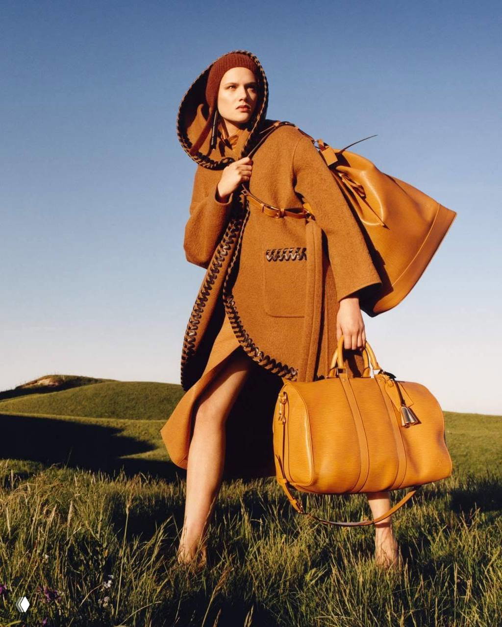 Модель в коричневом пальто держит крупную кожаную дорожную сумку Louis Vuitton на фоне скошенного поля и ясного неба.
