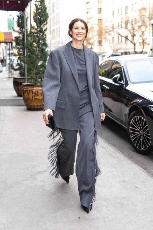 Уличный кадр: женщина в сдержанном тёмно-сером костюме с широким жакетом и бахромой на брюках, городской тротуар и автомобиль на заднем плане.