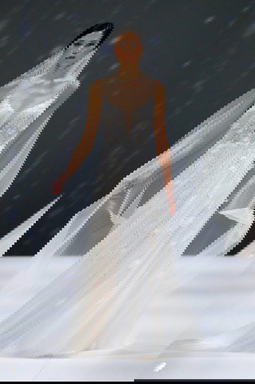 На подиуме модель в расшитом бисером и кристаллами свадебном платье с длинной фатой и тонкими бретелями, переливающийся декор.