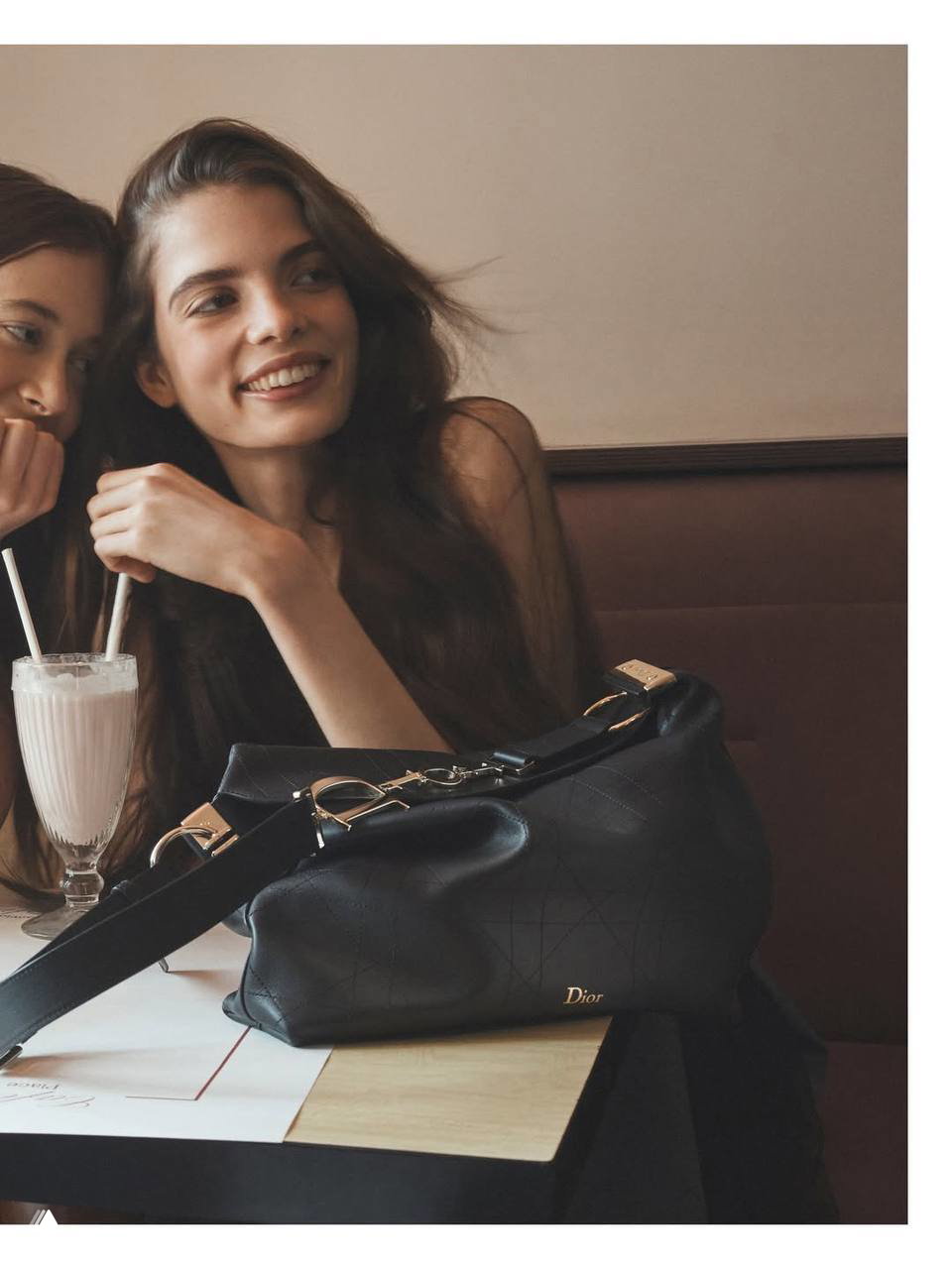Улыбчивая модель с темной сумкой Dior на столе и молочным коктейлем, тёплый повседневный вайб парижского утра и стильный аксессуар в кадре.
