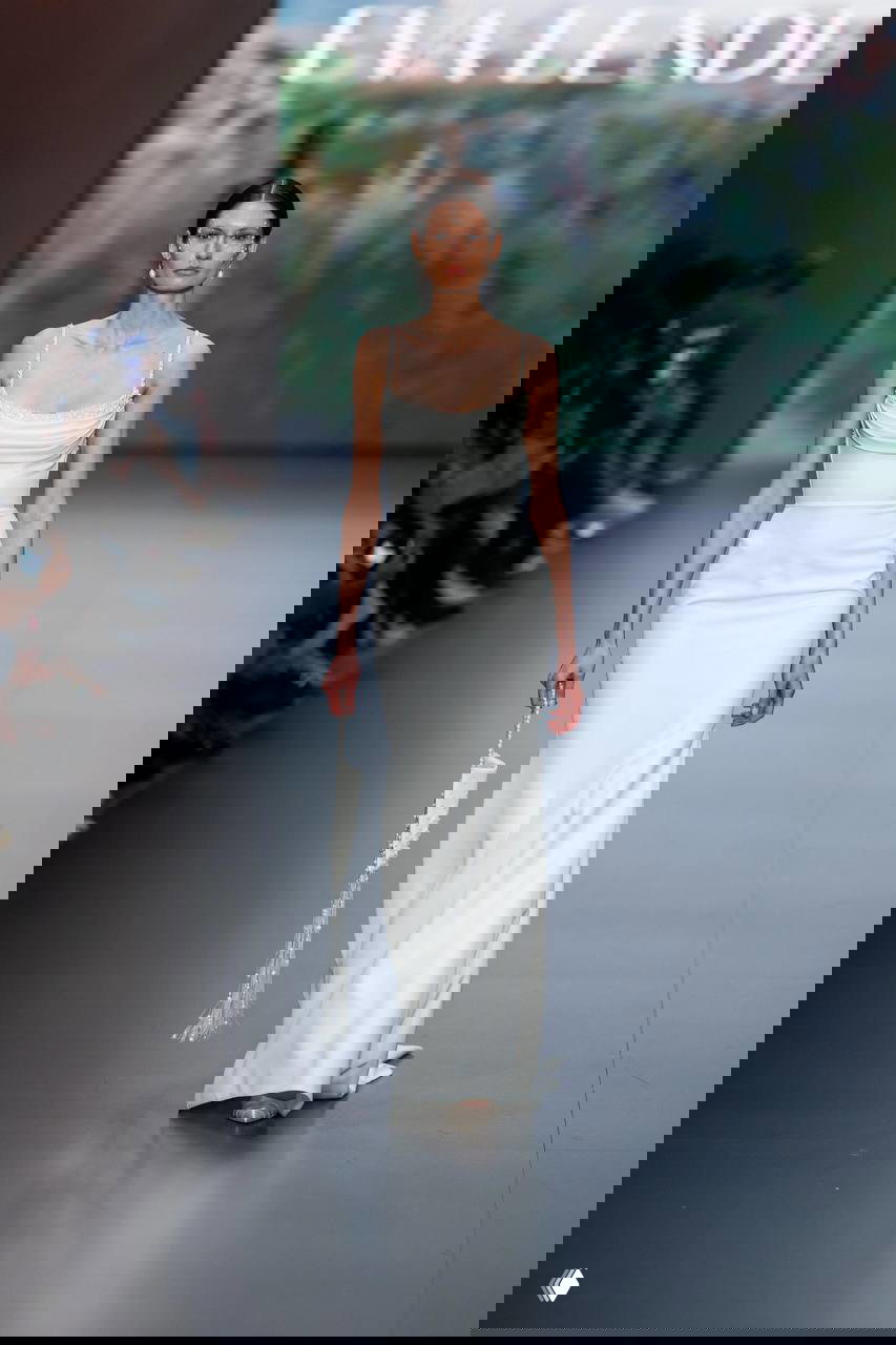 Стройная модель в узком колонном платье белого цвета с аккуратно драпированным верхом и небольшим аксессуаром в руке на показе.