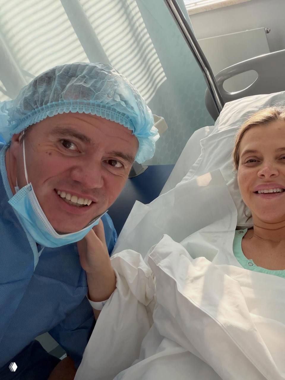 Селфи из палаты: улыбающаяся Марина лежит в больничной постели рядом с мужчиной в медицинском чепчике и маске, в кадре виден интерьер родильного отделения.