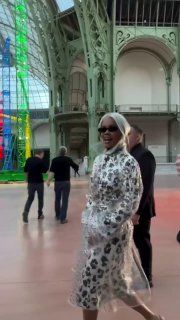 Дженни из Blackpink в прозрачном принтованном плаще и очках на фоне стеклянного павильона и инсталляции во время показа Chanel в Париже.