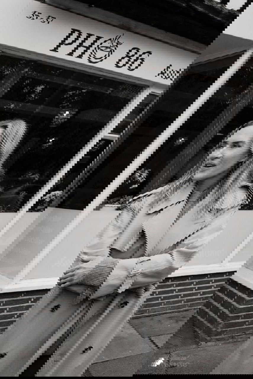 Актриса стоит перед витриной кафе в овчинном плаще — чёрно‑белый снимок с городской витриной и вывеской позади.