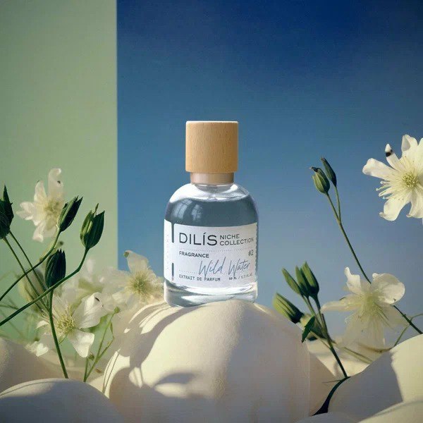 Флакон Dilis — Wild Water среди белых цветков на пастельном фоне; изображение подчёркивает свежий старт с лимоном и можжевельником и хвойно-амбровую базу композиции.