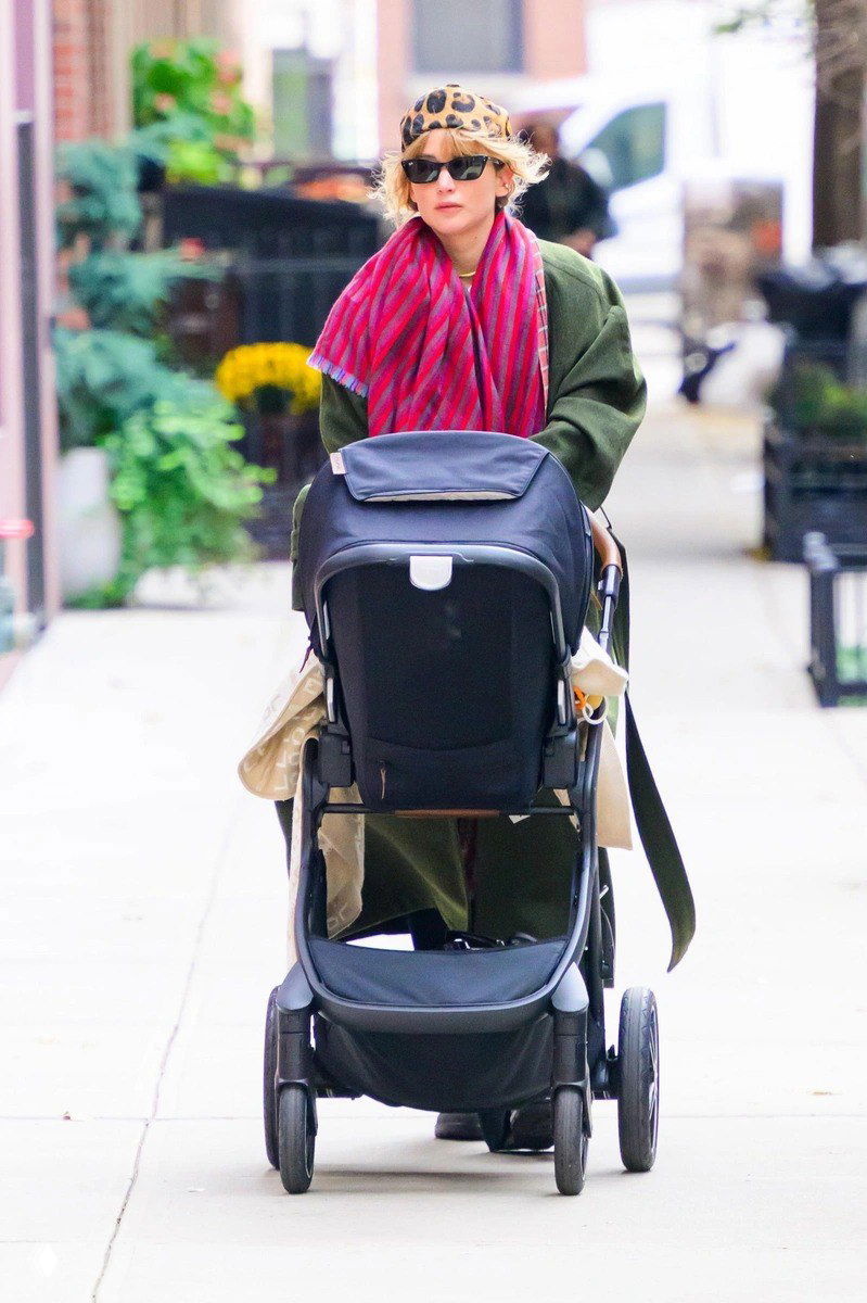 Женщина в тёмном оверсайз-пальто и леопардовом платке, толкает детскую коляску; уличный стритстайл-кадр с городским фоном и деревьями.