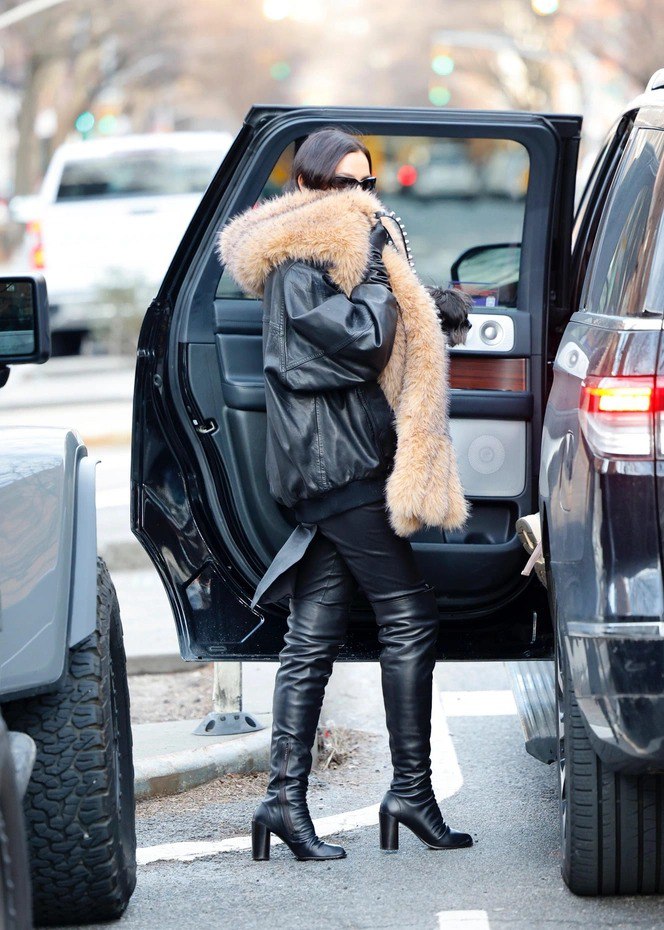 Городская сцена: тотал-блэк образ с объёмным светлым меховым шарфом, крупным пальто и высокими ботфортами на асфальте.