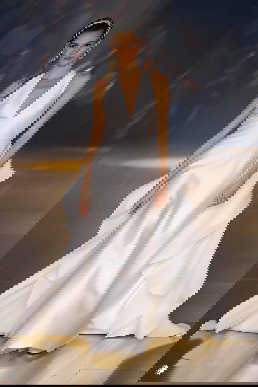 Платье из плотного атласа с рюшами на юбке и выразительным объемным шлейфом — модель идет по подиуму в свете софитов.