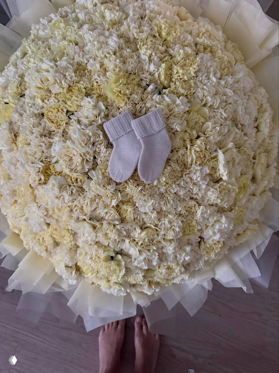 Крупный план большой круглой композиции из светлых бело‑жёлтых цветов, по центру положены маленькие вязаные носочки для малыша как символ подарка и торжества.