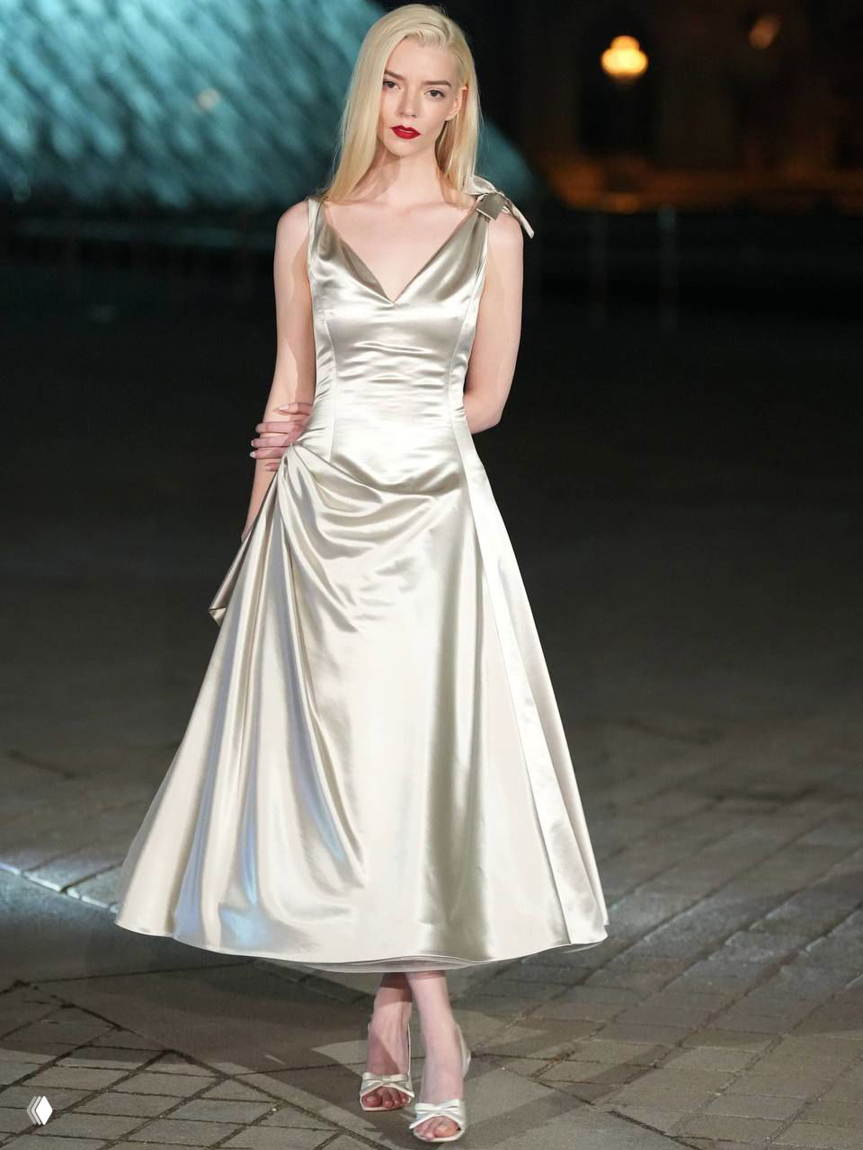 Женщина в атласном светлом платье-миди позирует у стеклянной пирамиды Лувра вечером, акцент на силуэте и блеске ткани